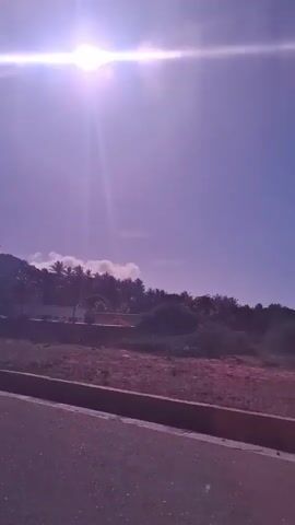 The video displays a bright, sunny daytime scene. The sun is high in the sky, creating a strong glare and lens flare. The sky is a clear blue with some distant, faint clouds visible near the horizon. A prominent hillside covered with abundant palm trees and other green vegetation dominates the background, suggesting a tropical or coastal environment. Several small buildings are nestled among the trees at the base of the hill. In the foreground, there is a paved road or highway, featuring a raised curb along its side. Adjacent to the curb is a strip of dry, reddish-brown earth. The perspective suggests movement along the road, likely from a vehicle. No people or specific activities are discernible. The intense sunlight indicates it is likely midday or early afternoon. The provided context states the location is Playa Parguito, Venezuela.