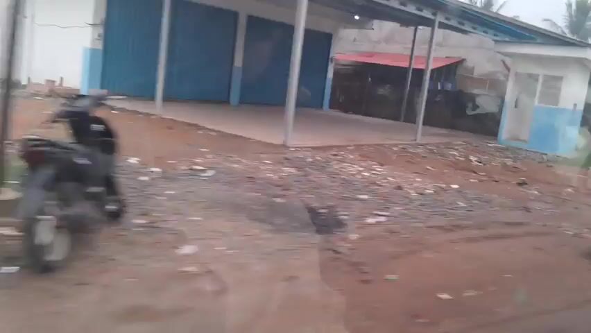 The video shows a series of brief, shaky shots taken from a moving vehicle, likely a car. The environment appears to be a rural or semi-urban area, characterized by dirt roads, occasional puddles, and an accumulation of scattered debris, including plastic bags and other trash. Buildings visible in the shots are simple and appear to be of modest construction. In one frame, a small, light blue and white booth-like structure is visible. Another frame shows a low-lying building with a red roof. Later frames reveal residential-style houses, some with grassy areas and trees. Objects such as plastic containers and what appear to be fuel canisters are scattered outside some of the buildings. The video is unclear and the camera movement suggests it was filmed casually, possibly as a personal record or while traveling. There are no clear interactions or actions involving people, as any individuals are either too blurry to identify or are only briefly glimpsed. The overall impression is of a landscape that is somewhat unkempt and possibly experiencing a period of wet weather, given the presence of puddles.