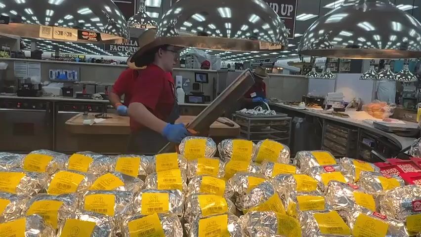 The scene shows an indoor food service area in a large store, likely a Buc-ee's, given the branding on some items. The primary focus is on a counter filled with dozens of individually wrapped sandwiches, each in foil and marked with a yellow label. Behind the counter, three employees wearing red shirts and cowboy hats are preparing food. One employee is slicing a large piece of meat on a cutting board. The other two are working on other tasks. Overhead are several large, shiny pendant lights reflecting the activity below. Signage is visible above and around the counter, including "Tortilla Factory" and promotional materials for various food items. The environment suggests a busy, commercial food preparation area within a larger retail space.