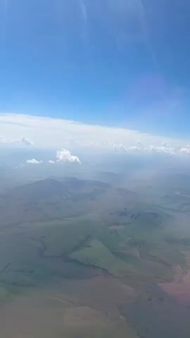 This is an aerial view from an airplane window looking down at a vast landscape under a bright blue sky with scattered white clouds. The terrain below is a mix of rolling green hills and flatter, possibly cultivated fields, with some areas appearing in shades of brown and pink. Sunlight streams down, creating visible rays through the atmospheric haze. The perspective suggests the plane is at a considerable altitude, offering a panoramic view of the earth's surface. There are no visible people or man-made structures, except for what might be distant roads or field divisions. The overall impression is one of natural, undeveloped or lightly developed, open space.