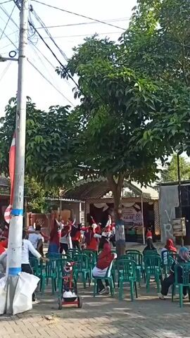 The video shows a group of people, mostly dressed in red, participating in an outdoor exercise routine. They are arranged in rows on a paved surface, with some standing and others seated in green plastic chairs. A tall pole with electrical wires and a flag is prominent in the foreground. In the background, there is a stage with a banner and some onlookers. The overall atmosphere appears to be one of community activity or an organized event.