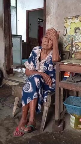 An elderly woman, dressed in a blue and white patterned dress, sits on a rustic wooden bench, her head propped up with her hand in a pensive pose. She wears a pair of rubber sandals.  She is positioned next to a similarly aged wooden table cluttered with miscellaneous items. The setting is an older, possibly weathered space, with worn walls and flooring. A doorway behind her reveals another room, where another elderly woman, wearing a similar dress, enters carrying a tray.