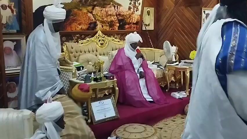 The video depicts a scene in a room with wood paneling. The room is decorated with multiple framed photographs on the walls. A central figure, likely an elder or leader, sits on an ornate, gold-trimmed sofa. This person is wearing a white turban and a pink robe and is speaking into a microphone. In front of the sofa, a small gold table holds various items, including what appears to be trophies and a framed certificate. Several individuals wearing traditional white robes and turbans are present. Some are standing and appear to be observing the central figure. The room's decor suggests a traditional or cultural setting, possibly associated with royalty or leadership.