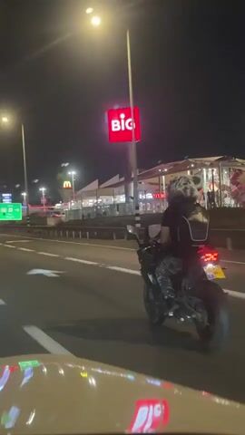 A motorcycle with a rider wearing a helmet and camouflage pants rides down a highway at night. The rider has a backpack on the back of the motorcycle. In the background, there is a large red sign with white lettering that says "BIG". There are also streetlights, buildings, and other cars on the highway. The motorcycle has its headlights on and the taillight is visible. The video is shot from a moving vehicle.