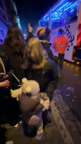 A group of people are standing and dancing on a street at night. A woman in a black jacket kneels holding a small child wearing a hooded snowsuit. Other women stand nearby, dancing and looking towards a truck that is partially visible behind them. The truck has blue lights and decorations. A man in a bright orange safety vest stands near the back of the truck. Confetti litters the ground.