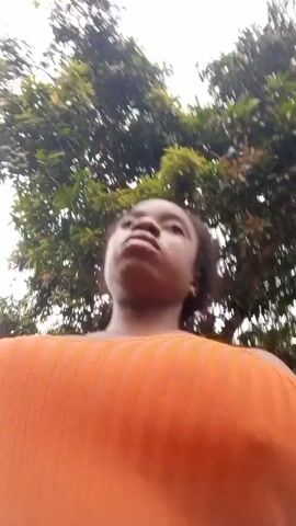 A woman with brown skin and dark hair is recording a selfie video while standing outdoors. She is wearing an orange ribbed top.  The camera is pointed up at her, showing the sky and treetops in the background. She looks up and then down at the camera. The camera moves slightly as she holds it. The sky is overcast.