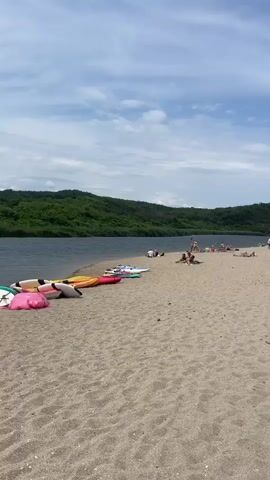 The video shows a wide sandy beach with several people scattered across it, some lying down and others standing. In the foreground, on the left side of the frame, are several kayaks and paddleboards lined up on the sand. To the left of the beach is a body of water, possibly a lake or a calm river, with green trees and hills in the background. The sky is blue with scattered white clouds. Further down the beach, there are more people and some structures, including a sailboat and a small building. There is also a piece of driftwood lying on the sand. The overall scene depicts a peaceful day at the beach with various recreational activities.
