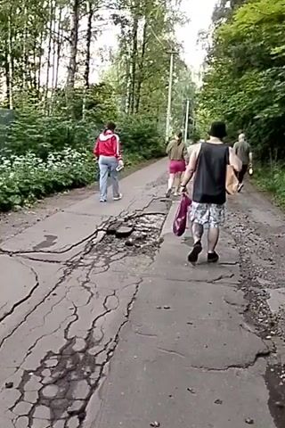 A group of four people are walking down a cracked and damaged asphalt road that appears to be in a wooded area. The road is uneven and has large potholes. The people are walking away from the camera and are spaced out along the road. The first person is wearing a red jacket and blue jeans. The second person is wearing a pink skirt and a green top. The third person is wearing a black tank top and patterned shorts, carrying a red bag. The fourth person is in the distance and is wearing lighter clothing. The trees on either side of the road are lush and green, suggesting it is summer. The sky is overcast.