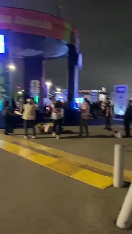 This media file captures an outdoor scene at night, likely a public event or gathering. The setting appears to be an urban environment, possibly near a venue or entertainment space, suggested by the illuminated structures and banners. Several people are visible, engaged in various activities. In the foreground, a person in a striped shirt is dancing or moving energetically. To their left, other individuals are also present, some appearing to socialize or observe. In the background, there are more people, as well as what seem to be booths or displays with bright, colorful lighting, including purple and green. A large archway with illuminated signage is prominent in the left portion of the frame, with what appears to be "LOS ANGELES" visible on it. There are also white vertical posts in the foreground, possibly part of a barricade or traffic control. The overall atmosphere seems lively, with people enjoying themselves in a festive environment. The time of day is clearly nighttime, and the weather appears to be clear. The presence of FIFA World Cup branding suggests the event might be related to the 2026 World Cup, which is scheduled to be hosted in Los Angeles among other cities.