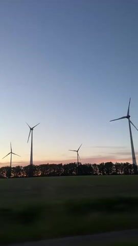 The video shows a view of a field with wind turbines at dusk. The camera is moving, likely from a moving vehicle. The turbines are silhouetted against the evening sky, which has hues of orange and blue. A row of trees separates the turbines from the field. The overall impression is one of motion and a rural landscape at twilight.
