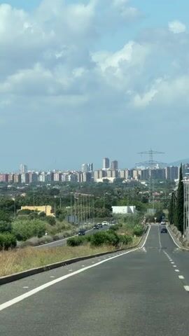 The video shows a long shot of an empty two-lane road in a rural area, with a city in the distance. The road has a solid white line on the right shoulder and a broken white line separating the two lanes. The left side of the road is bordered by a grassy area and trees, while the right side has a row of streetlights and tall, thin trees. In the distance, a city skyline is visible with numerous buildings and a few tall skyscrapers. The sky is partly cloudy with some blue patches visible. A high-voltage power line structure is also visible on the right side of the city.
