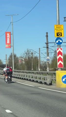 Three cyclists are riding on a bridge on a clear, sunny day. They are all wearing helmets and cycling gear. The first cyclist is wearing a white helmet and a light gray shirt, while the second and third cyclists are wearing red helmets and red shirts. A red banner with the word "ПОБЕДА!" (Victory!) is on a lamppost near the bridge. Overhead electrical lines are above the cyclists. Beyond the bridge, a sparse tree line borders what looks like a railway area.