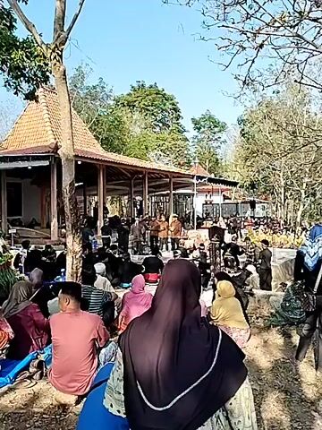The video shows an outdoor gathering of people. In the foreground, several individuals are seated on blue tarps, facing a building with a tiled, pyramidal roof and an open-air veranda. A tall, elaborate floral arrangement or offering stands in front of the building. A crowd of people are visible on the tarps and around the building. Some individuals are dressed in traditional attire. The environment appears to be rural, with trees and a clear sky. The activity suggests a religious or cultural ceremony.