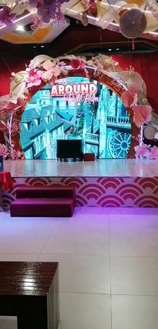 The video shows a stage decorated with a large, circular screen displaying images of a city. The words "AROUND Viet NAM" are displayed above the image. The stage is adorned with pink flowers and fabric, and there are red steps leading up to the stage. There is also a small table covered with a red cloth on the left side of the stage. The floor in front of the stage is tiled.