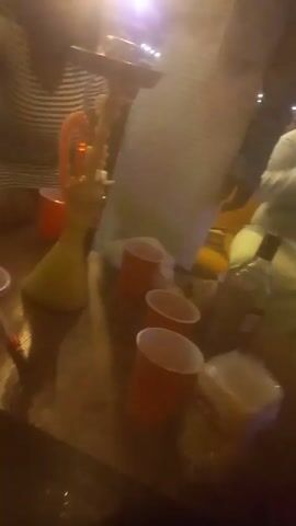 The video appears to be taken in an outdoor lounge or bar area, possibly at night, judging by the dim lighting. Multiple people are seated at tables, with various drinks and what seems to be a hookah present. There are several red plastic cups on the tables, suggesting a casual gathering. The environment includes outdoor seating, tables, and ambient lighting. The camera moves around a little bit as the person recording tries to get a clearer shot of the scene. The focus isn't very sharp and the picture quality is not the best, but it gives a general impression of people relaxing and socializing in a relaxed setting.
