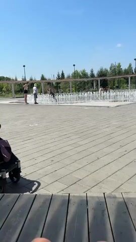 A bright, sunny day is depicted with a clear blue sky. The foreground shows the legs of an individual sitting, with their feet visible and wearing sandals. The midground features a paved area with interlocking gray tiles. Beyond this, a large, circular splash pad with numerous water jets is active, with several children playing in the water. In the background, a row of green trees and some structures are visible, including covered walkways supported by posts. Two individuals, a woman and a child, are standing to the left of the splash pad. The overall scene appears to be a public park or recreational area.