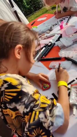 The video shows a close-up of a young girl with dark hair wearing a yellow and black patterned shirt and a yellow wristband. She is sitting at a table covered with various craft supplies, including markers, glue, and small containers filled with beads or rhinestones. The girl is focused on her activity, holding a small object in her left hand and a pen or marker in her right, appearing to be decorating the object. In the background, another person, possibly a child, is also visible at the table, engaged in crafting. The setting appears to be outdoors, with natural light and greenery suggested in the background. The overall scene depicts a crafting or art activity for children.
