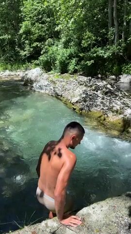 A young man is partially submerged in a shallow, clear river and is preparing to jump in. He is wearing light-colored underwear. The river is surrounded by lush green trees and rocky banks. Sunlight filters through the trees, casting shadows on the water and the man's body. The water appears to be flowing rapidly over rocks.
