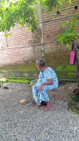 An elderly woman sits on a makeshift swing outdoors, suspended from a tree branch. She wears a blue and white patterned pajama set and red sandals. Her hair is pulled back in a bun. A brick wall runs along the back of the scene, with patches of moss at its base. The ground is covered in small gravel. A pink handbag hangs from the tree next to her. A child's head is visible in the foreground.