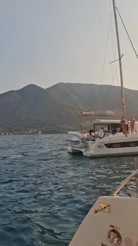 The video shows a white catamaran named "BALI" with several people on board sailing on the water. The catamaran has a tall mast and appears to be moving. In the background, there are green mountains and a clear sky. The water is choppy, and there is another boat in the foreground, partially visible. The lighting suggests it is daytime, possibly late afternoon or early morning.