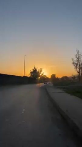 The video shows a road at sunset. The sun is setting directly ahead, creating a golden glow. There are trees and bushes on either side of the road, and a wall on the left. Cars drive on the road. The sky is a gradient of blue to orange. The road appears to be wet.