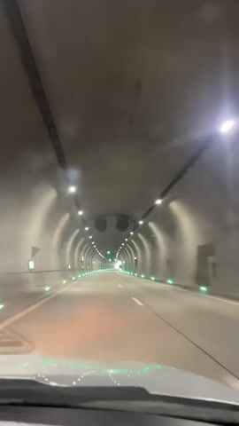 The video shows a first-person perspective of driving through a tunnel. The tunnel has a curved ceiling and concrete walls. There are lights lining the ceiling, and green lights are embedded along the lower parts of the walls. Traffic signs are visible on the walls. The road is paved and marked with white lines. The video is short and does not show any people or other vehicles clearly.
