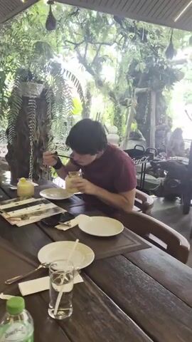 The video shows a young person with dark hair wearing a maroon t-shirt eating from a yellow container with a spoon. They are seated at a wooden table in an open-air restaurant with a brick pillar on the left. Lush green foliage hangs from above, suggesting an outdoor or semi-outdoor setting with a natural, tropical ambiance. A pond with some rocks is visible in the background. On the table, there are plates, cutlery, a glass of water with a straw, and what appears to be a menu or a stack of papers.

Later in the video, a woman with dark curly hair wearing a black spaghetti strap dress joins the table and also picks up a yellow drink with a straw, looking at it intently. She then proceeds to drink from it using the straw. The setting appears to be a restaurant in Sakhu, Thailand, as indicated by the provided location context. The overall atmosphere seems relaxed and casual, possibly during daylight hours given the bright, natural lighting. Visible text includes a "Coca-Cola" sign in the background and what looks like writing on the menu or papers on the table, but it is not clearly legible.