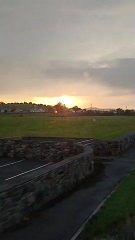The video shows a blurry, side-view of a rural landscape at sunset. The sun is low on the horizon, casting a warm glow across the sky, which is filled with scattered clouds. In the foreground, there is a stone wall bordering a paved path or road. Beyond the wall, a wide, green field stretches out, dotted with a few scattered trees and distant houses. There is a metal gate visible on the left side of the frame. The camera appears to be moving, likely from a vehicle, as indicated by the motion blur.