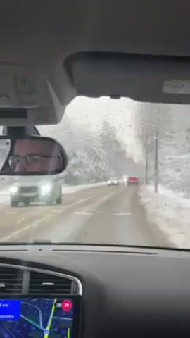 The video shows a car's point of view driving on a snowy road. The road is covered in slush and snow, and the trees on either side are laden with snow. There are other cars ahead on the road. The car's dashboard is visible, showing the speedometer and a navigation system. The rearview mirror reflects the face of a person wearing glasses. The weather appears to be overcast, and it might be snowing or raining.