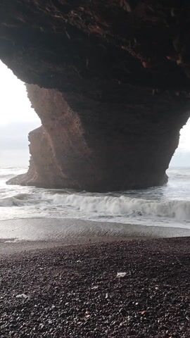 The video shows a dark-colored rocky beach under the shadow of an overhanging rock formation, facing a sea. The beach is composed of small, dark pebbles. Waves crash around a large, reddish-brown rock formation standing in the water, further out from the shore. The sky is overcast and grayish.