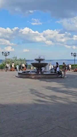 A fountain is in the center of a paved area. People are scattered around the fountain, some sitting on the edge and others standing. In the background, there is a body of water with boats. The sky is blue with white clouds. There are lampposts on either side of the scene and trees to the left.