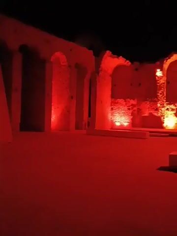 The video shows an outdoor scene at night, illuminated by red lights. The foreground is a flat, red-lit surface, likely the ground. In the background, there are stone ruins with arched openings. The structures appear ancient and possibly part of a historical site. The red lighting creates a dramatic and somewhat ominous atmosphere. No people or specific actions are visible in the frame.