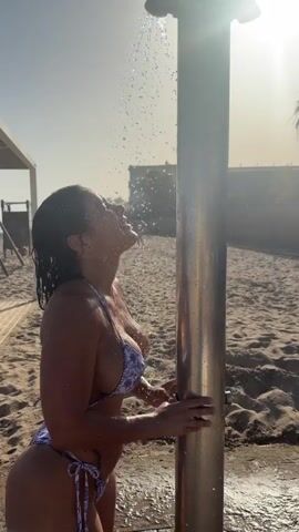 A person wearing a purple and white patterned bikini is standing under an outdoor shower on a sandy beach. The sun is shining brightly, creating a lens flare effect. The person is rinsing their face and hair with water. In the background, there are beach chairs and palm trees.