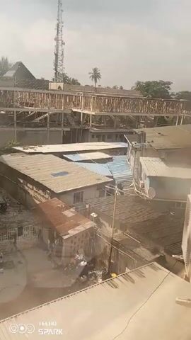 Here is a description of the video:
The video is a high-angle shot taken outdoors, providing a view of a densely populated urban area. In the foreground and midground are a cluster of buildings with diverse roof types, including corrugated metal and flat surfaces. These buildings appear to be residential or small commercial structures. In the background, a multi-story building under construction is visible. The framework of the building is exposed, revealing the structure's skeleton. A telecommunications tower rises above the other structures in the vicinity. The sky is overcast, with muted sunlight filtering through the clouds. In the center of the frame, a man is performing some kind of exercise on a paved surface between buildings. Various objects and vehicles are scattered around the area, adding to the cluttered scene. The camera is stationary. The overall impression is of a bustling, densely populated area with ongoing construction and everyday activities.
