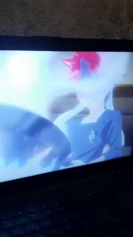 The video shows a close-up of a laptop screen displaying an anime scene. The scene depicts a character with red hair and a blue uniform, sitting and reaching out towards something off-screen. There is a bright, glowing blue and white aura surrounding the character and their extended arm. The background is blurred but suggests an indoor environment. The scene then transitions to a colorful, abstract display of swirling purple and white light with small, shimmering particles.