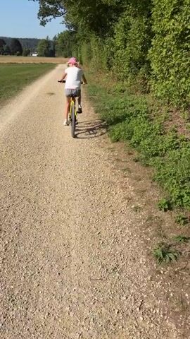 A girl wearing a pink hat and white t-shirt rides a bicycle on a dirt path. The path is lined with trees on the right side and a field on the left. The sky is clear and blue. In the distance, there are hills and some buildings. The camera is following behind the girl, capturing her ride. At 00:04, another bicycle enters the frame from the left, casting a long shadow on the path.