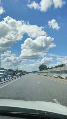 The video is a first-person view from a vehicle traveling on a highway. The sky is blue with numerous fluffy white clouds. The road is asphalt with white lane markings. On the left side of the road, there is a metal guardrail. On the right side of the road, there is a white sound barrier and some trees. There are streetlights visible along the highway. Other vehicles, including a truck, are visible in the distance on the highway. The sun is shining, casting shadows.