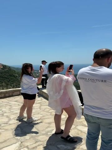 A medium shot shows a man in a white t-shirt and jeans holding a camera and taking a photo of a couple at a wedding. The man is standing on a stone patio overlooking the ocean. In the foreground, a woman in a white t-shirt and shorts is taking a photo with her phone. Next to her, another woman in a pink robe and shorts is holding a white veil. In the background, the bride in a white wedding dress and the groom in a dark suit are walking down a set of stone stairs towards the ocean. The sky is blue and clear.