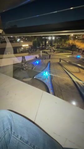 This media file captures a nighttime scene, likely in Nicosia, Cyprus. The viewpoint is from a slightly elevated position, looking down and outwards onto an open public space.

The primary subjects are the legs of a person in blue jeans and sneakers, positioned in the foreground, suggesting they are sitting or resting on a ledge. In the background, the scene unfolds across a paved area with geometric landscaping and water features. These water features are illuminated with vibrant blue lights, creating a dynamic and modern aesthetic.

Several people are visible as small figures in the distance, walking or standing in the illuminated plaza, indicating it's an active public space despite the late hour. The background also shows buildings with illuminated windows and signage, hinting at shops or restaurants, one of which has visible text that appears to read "CAFFE." The overall lighting suggests it is nighttime. The sky above is dark, with a faint suggestion of light pollution.

The presence of the illuminated water features and the pedestrian activity point to an urban environment, possibly a recently developed public square or promenade. The mood evoked is one of relaxed observation of an evening cityscape. The media file appears to be a personal perspective, capturing a moment of leisure in a well-lit, modern urban setting.