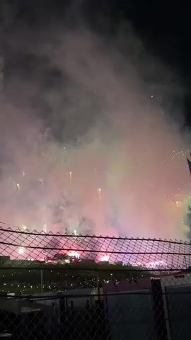 This video captures a nighttime fireworks display, showcasing vibrant colors and plumes of smoke against a dark sky. The foreground is partially obscured by a dark wire fence, suggesting the viewer is at a distance from the event. Beyond the fence, a large gathering appears to be enjoying the spectacle, with various lights illuminating the ground. A tall blue flag is visible on the right side of the frame, adding a structural element to the scene. The fireworks burst in the air, creating a dynamic and colorful display, with smoke gradually filling the sky.