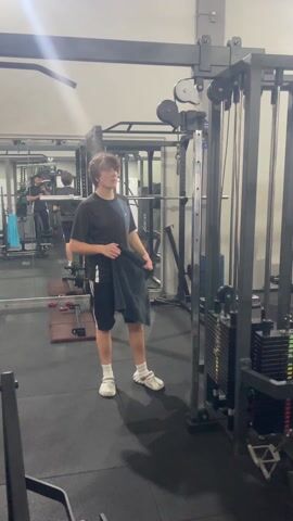A young male in athletic gear is standing in a gym, holding a towel. He glances towards the camera, smiles, and wipes his forehead as he begins to walk. He is wearing a black t-shirt with blue and white stripes, black athletic shorts with white stripes, white socks, and white shoes. He is standing next to a cable weight machine in a gym with mirrors lining one wall. In the mirrors, another person can be seen filming with a camera. Other weightlifting equipment is visible in the background. The floor is black.