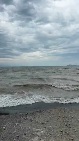 The video shows a beach with gray, choppy water under a cloudy sky. The shore is made up of dark sand and pebbles. In the middle of the video, a young man with short, dark hair walks from right to left across the screen. He is wearing a white t-shirt with a black design on the sleeve and black pants. He is walking along the edge of the water, with waves breaking on the shore. The man is the only person visible in the video.