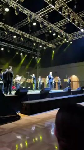 Here is a description of the video:

A band is performing on an outdoor stage. The band consists of a singer in a dark suit and several musicians playing brass instruments. The stage is illuminated by bright white and green spotlights. Above the stage is a complex metal structure with various lights. In the foreground, an audience member is visible. Another person in a jacket and light pants walks around on stage, taking photos. The setting appears to be an outdoor event at night, with a building with arched windows in the background.