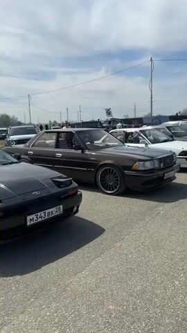 The video shows an outdoor car show on a sunny day. Various older cars are on display including a black Toyota Supra, a brown Toyota Crown with aftermarket wheels, and a red Lada with a Soviet flag. People are standing around in groups, talking and looking at the cars. The background includes power lines and light poles. The ground is paved with asphalt.