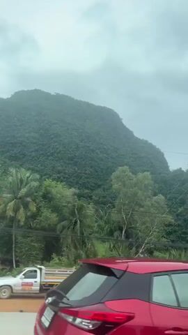 This is a short, vertical video clip captured from inside a vehicle, likely in Phang-nga, Thailand. The camera is pointed outwards, offering a view of a lush, green mountainside covered in dense vegetation. The terrain is characterized by steep, karstic rock formations, with visible white rock faces peeking through the greenery, hinting at the presence of caves or limestone cliffs.

In the foreground, the side and rear-view mirror of a red vehicle are visible, framing the scene. A road, with a reddish-brown dirt shoulder and a paved section with white markings, runs alongside the mountains. Power lines and a utility pole are prominent features, suggesting the presence of infrastructure in this natural landscape.

The vegetation is a mix of tall trees and shorter, broad-leafed plants, possibly palms or similar tropical foliage. The sky is overcast, indicating cloudy or possibly rainy weather. The overall impression is one of natural beauty and a somewhat remote, undeveloped setting, likely during the daytime due to the ambient light. There are no people visible in the frame. The video captures a scenic drive through a mountainous, tropical region.