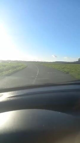 A car is driving down a narrow asphalt road through a rural area on a bright, sunny day. The sky is clear blue. On either side of the road are fields of green vegetation. The sun shines intensely into the camera through the windshield, causing glare. The camera view briefly shifts to show the rearview mirror and a wider view of the green field passing by outside the window.
