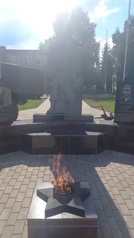 A video shows a memorial with a statue of a soldier holding a rifle, a lit eternal flame, and several plaques with images and names. A military truck is parked behind the memorial. The scene appears to be outdoors on a sunny day with a building and trees in the background.