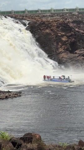 A large boat filled with people approaches a powerful waterfall in Guayana City, Venezuela. The boat, predominantly blue and white with a canopy, carries numerous individuals, many of whom are wearing orange life vests. Some passengers on the left side of the boat are standing and waving, while others sit towards the back. The waterfall cascades down a rocky, brown cliff face, creating a significant amount of white spray and turbulent water around the base. The boat is positioned within this turbulent area, with the strong current visible around it. The water in the foreground is dark gray. The scene suggests a tourist activity, offering an up-close view of the waterfall. Text, possibly "LAGOS," is partially visible on the side of the boat. The weather appears bright and clear, indicating daytime.