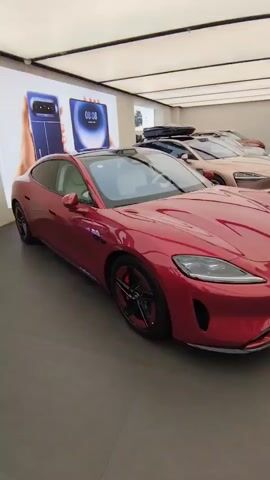 THE VIDEO SHOWS A RED SPORTS CAR PARKED INSIDE A SHOWROOM. THE CAR IS THE FOCUS OF THE VIDEO. THE CAR IS PARKED IN FRONT OF A LARGE DIGITAL SCREEN DISPLAYING AN IMAGE OF A BLUE FOLDABLE SMARTPHONE AND A CLOCK. THERE ARE OTHER CARS PARKED IN THE BACKGROUND. THE FLOOR IS DARK COLORED.