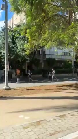 The video shows a street in what appears to be a residential neighborhood with apartment buildings and trees. A group of people wearing matching blue shirts and shorts are running on the street. Other people are walking on the sidewalk. The video seems to be taken from a moving vehicle. The weather is sunny.